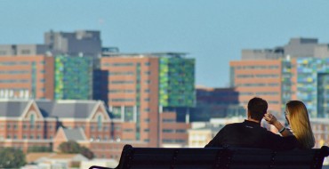 A woman and a man in their 30s on a park bench looking at a city on a date.