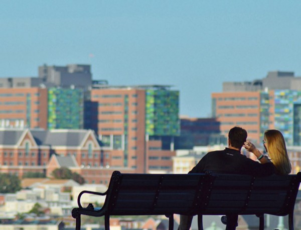 A woman and a man in their 30s on a park bench looking at a city on a date.