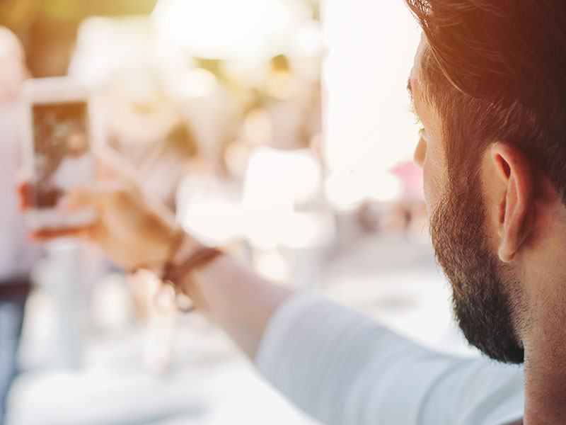 A man trying to take the best profile picture possible while standing outside and taking a selfie.