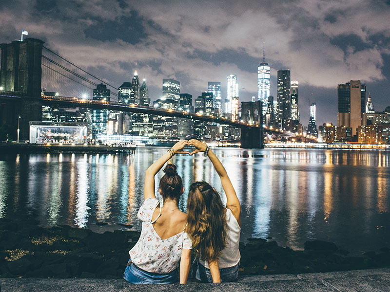 Two millennial girls sitting in front of a city scape on a dating, making a heart sign.