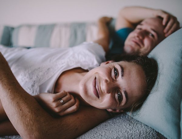 A woman who read the signs that he still loves you and is in bed smiling with her boyfriends arm around her.