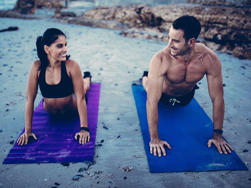 A happy couple doing yoga together on the beach and smiling at each other.