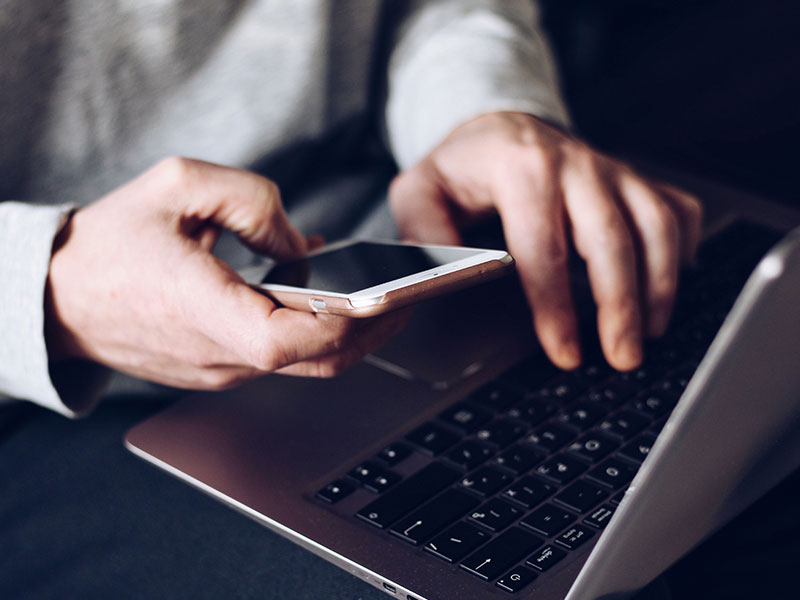 An online dating scammer typing at a computer while looking at a phone at the same time.