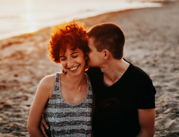 A woman who learned how to let your guard down, getting her cheek kissed on the beach while she laughs with her boyfriend.