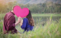 A couple getting creative in how to show affection by kissing behind a pink heart in a field.