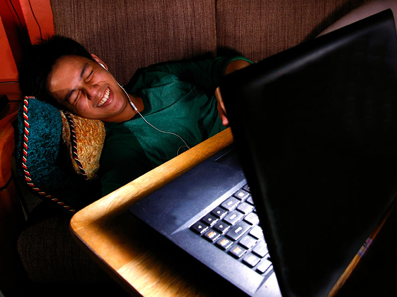 A man in an online relationships, laughing at his computer screen wrapped up in a blanket in bed.