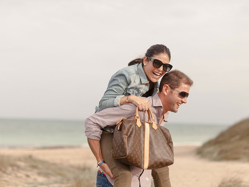 An older women dating a younger man getting a piggy back ride from her boyfriend and laughing on the beach.