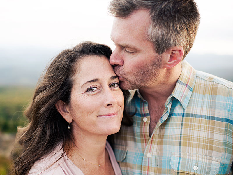 Two singles over 50, smiling on a beach and the man kisses the woman’s forehead.