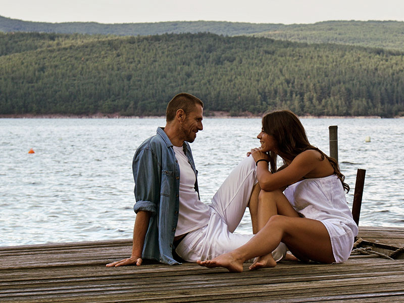 A couple who learned what to do on a first date, leaning in and talking to each other on a dock by a lake.