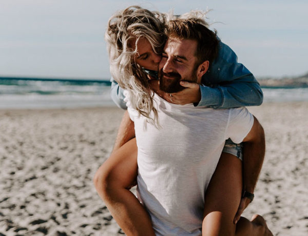 A woman who figured out what single men want, laughing and kissing her boyfriend’s cheek on the beach.