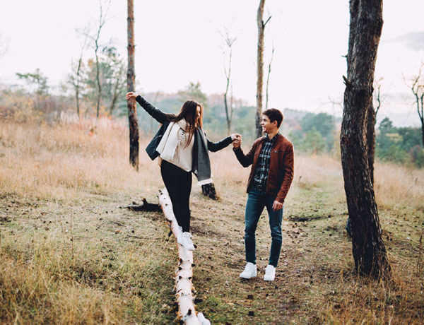 A girl who's wondering "is he thinking about me?" balancing on a log while the guy she likes helps her and looks on with a smile.