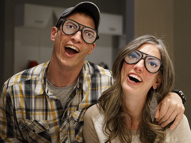 A woman who's dirty a nerdy guy, wearing nerd glasses with her boyfriend while they both make silly faces and laugh.