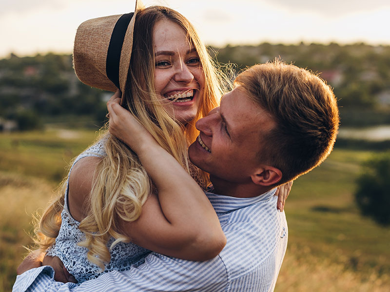 A couple who went through these stages of dating laughing as a man holds his girlfriend in the hair and she wears his hat.