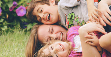A woman who's dating a single parent with full custody laughing as she wrestles in the grass with two kids.