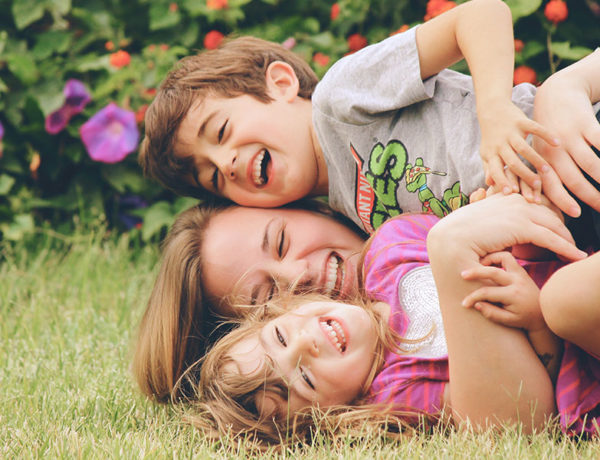 A woman who's dating a single parent with full custody laughing as she wrestles in the grass with two kids.