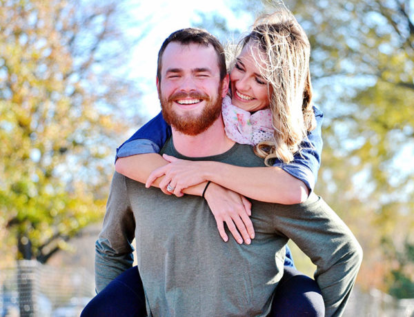 A man laughing as he carries his girlfriend on his shoulders as she looks at him and wonders, "how do men fall in love?"