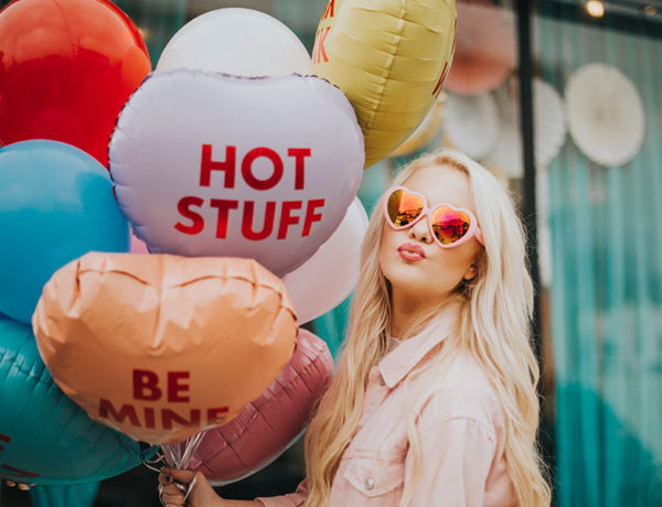 A girl with Valentine's Day balloons and heart sunglasses making a kissy face at the camera.