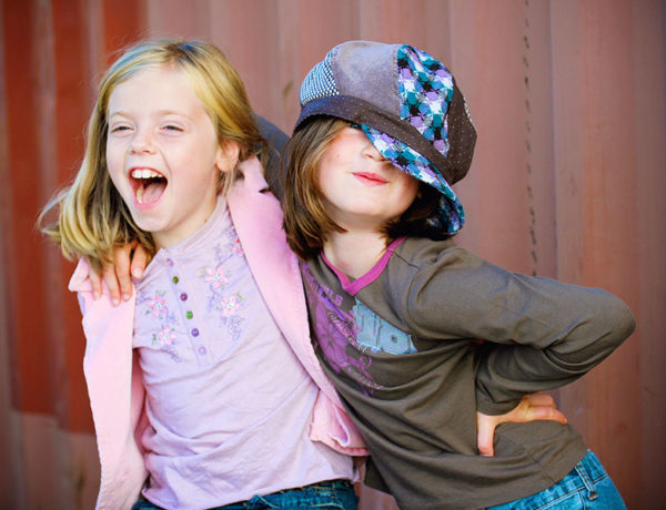 Two little girls who are childhood friends laughing and joking around together in front of a fence.