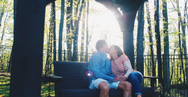 A couple who met on over 50 dating sites, kissing on a park bench as the sun shines through the trees.