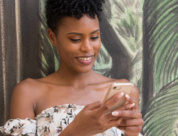 A woman wondering, "Should I text him first?" while looking into her phone outside.