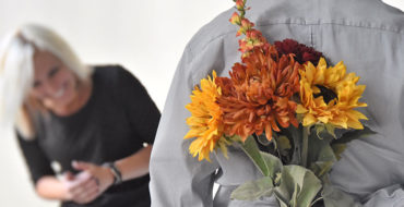 A man dating after 50, holding flowers behind his back while his date laughs in front of him.