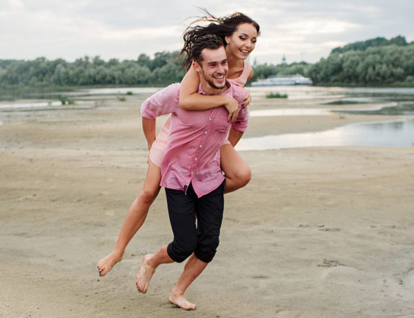 A girl wondering "does he like me" smiling and laughing as a guy friend carries her on his back on the beach.