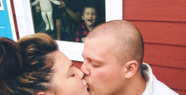 A woman who's a single parent and dating, kissing her date while her kids are inside pushing their faces against the window and making funny faces.