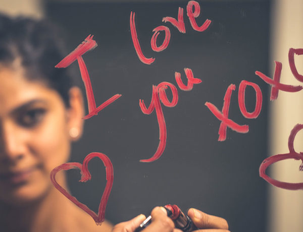 A woman writing, "I love you" on a mirror with lipstick, one of the romantic gestures for him that guys appreciate.