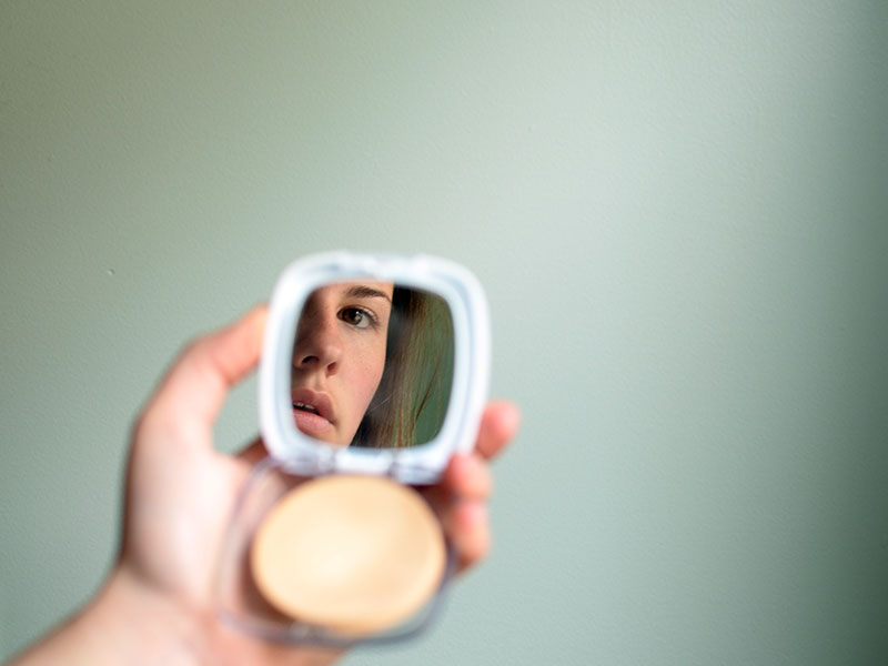 A woman who has body image issues, looking at her reflection in the mirror of her makeup compact.